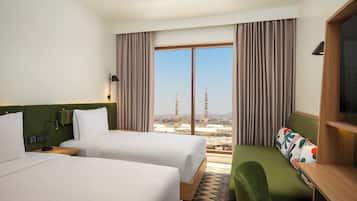 Habitación de hotel con dos camas, un sofá, vistas a un paisaje urbano con una torre, y un amplio ventanal con cortinas.
