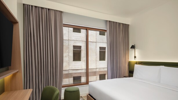 Quarto de hotel com cama, escrivaninha, cadeira, um banquinho, mesa de cabeceira e janela com cortinas.