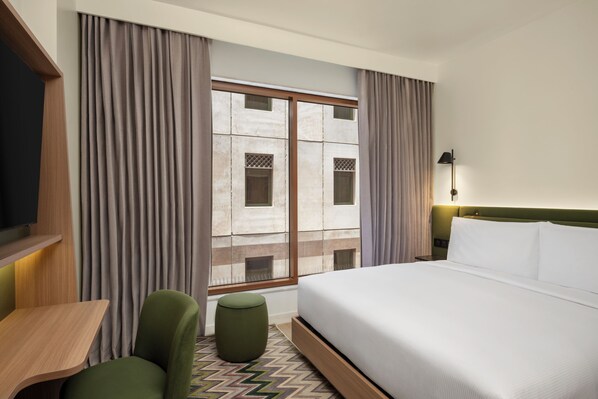 Ein Hotelzimmer mit Bett, Schreibtisch, Stuhl, Hocker, Nachttisch und einem Fenster mit Vorhängen.