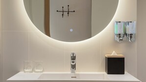 A modern bathroom with a round mirror, a sink, two glasses, a tissue box, and a hand sanitizer dispenser. - Hampton By Hilton Madinah Central District (Madinah)