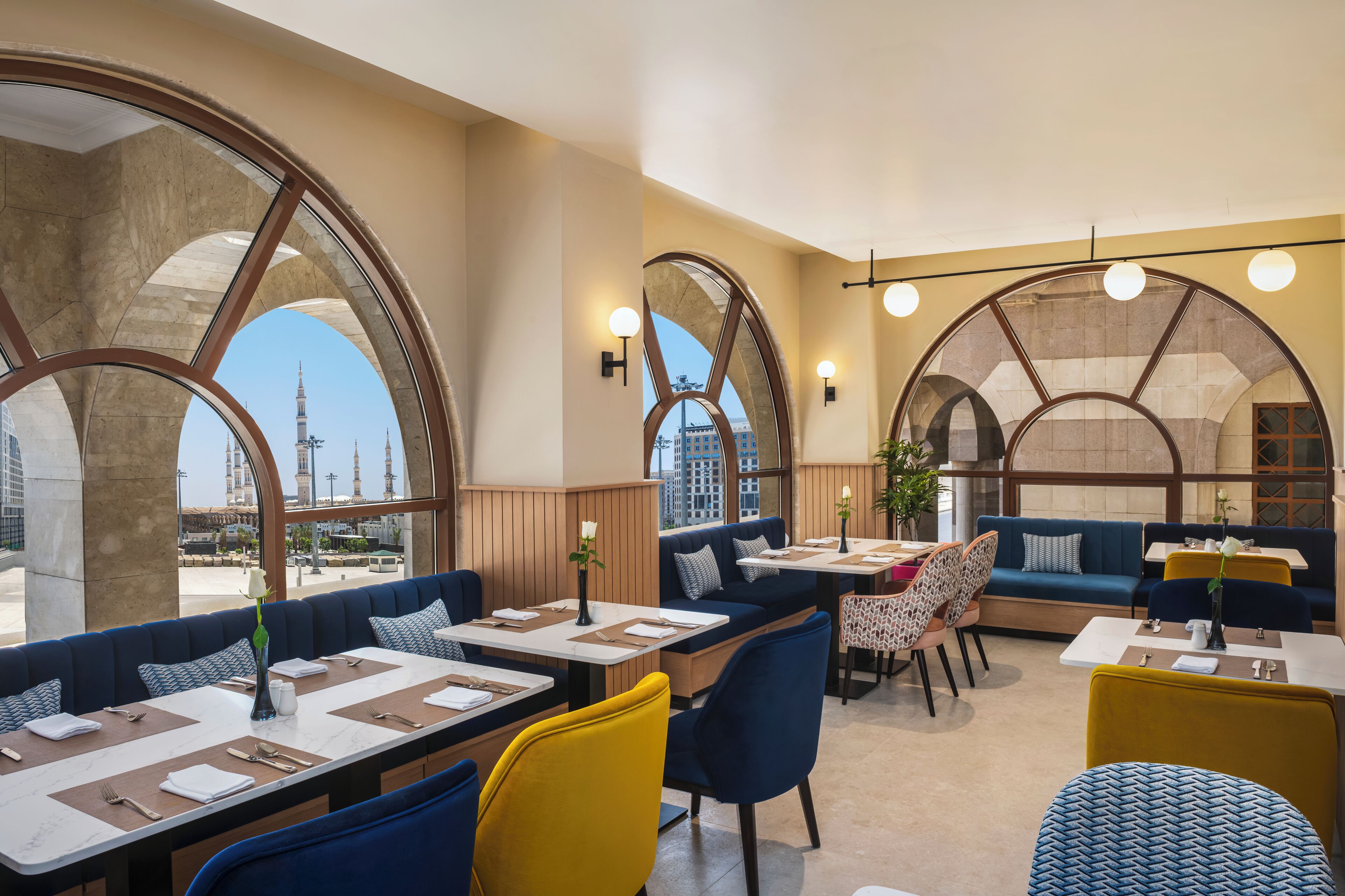 A restaurant with high arched windows, blue and yellow seating, and a view of a tower.
