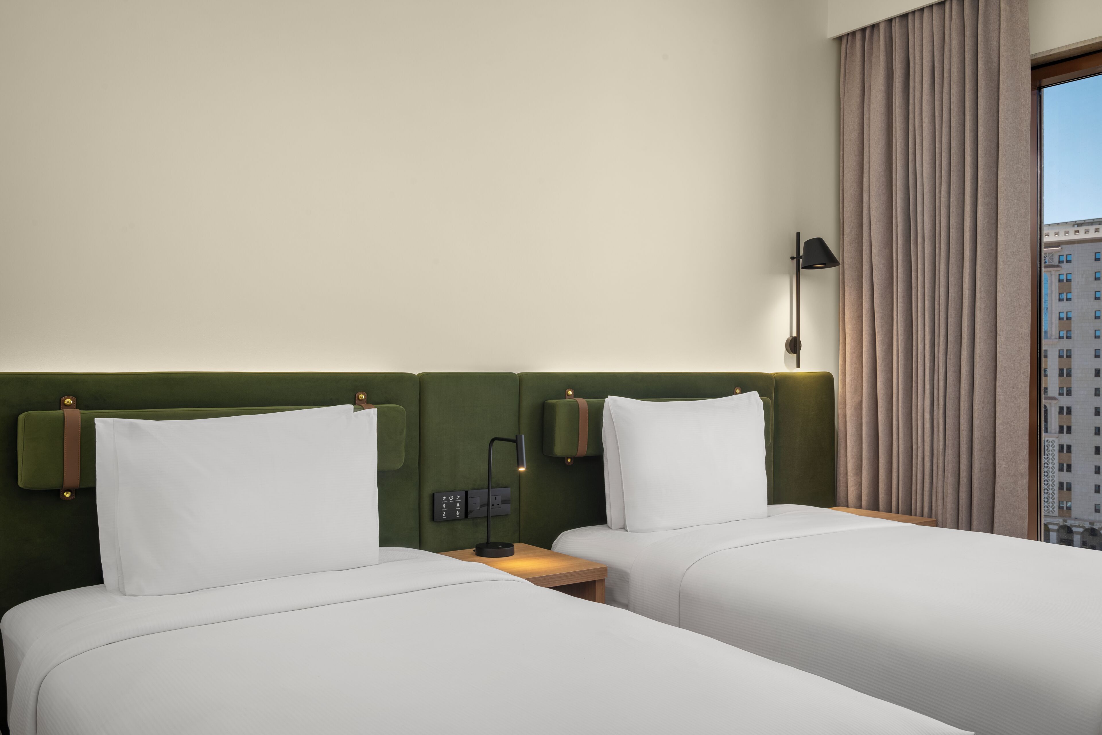 Una habitación de hotel con dos camas, un cabecero verde, una mesita de noche con lámpara y una ventana con cortinas.