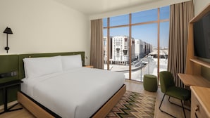 A hotel room with a large bed, a desk, a chair, a TV, and a view of the cityscape. - Hampton By Hilton Madinah Central District (Madinah)