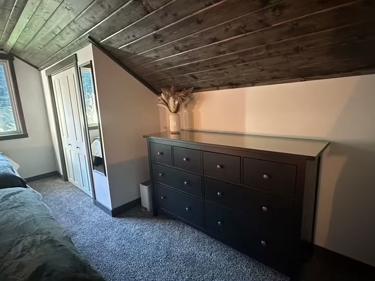 A bedroom with a sloped ceiling, a dresser, a window, and a door.