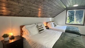Quarto com duas camas, uma mesinha de cabeceira com abajur, janela com vista para árvores e teto de madeira.