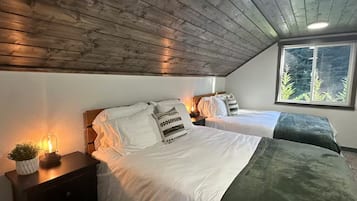 A bedroom with two beds, a nightstand with a lamp, a window with a view of trees, and a wooden ceiling.