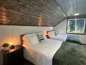Et soveværelse med to senge, et natbord med lampe, et vindue med udsigt til træer og et træloft.