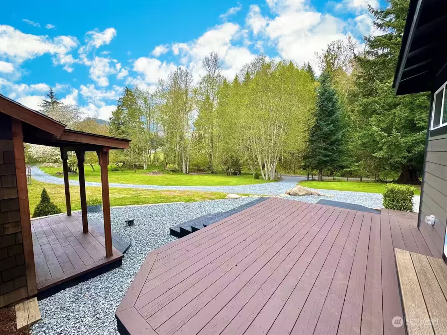 A wooden deck with a pergola, surrounded by a gravel path, trees, and a clear sky.