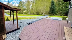 Um deck de madeira com pérgola, ladeado por um caminho de cascalho, árvores e céu claro.