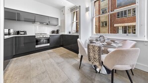 A modern kitchen with grey cabinets, a dining table with chairs, and a view of a brick building through large windows. - 3 -Berry Street EC1V (London)