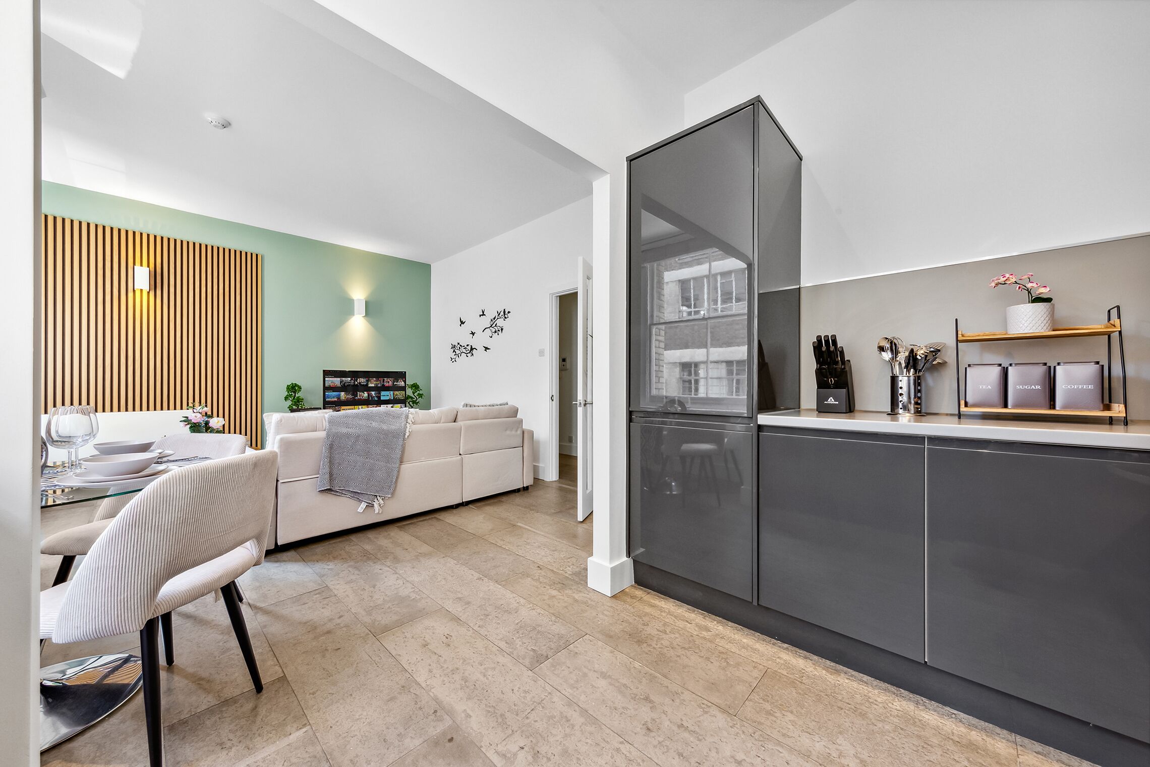 A modern kitchen with a built-in fridge, a dining area with a white table and chairs, and a living room with a sofa and a decorative wall.