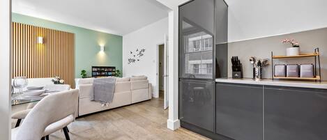 A modern kitchen with a built-in fridge, a dining area with a white table and chairs, and a living room with a sofa and a decorative wall.