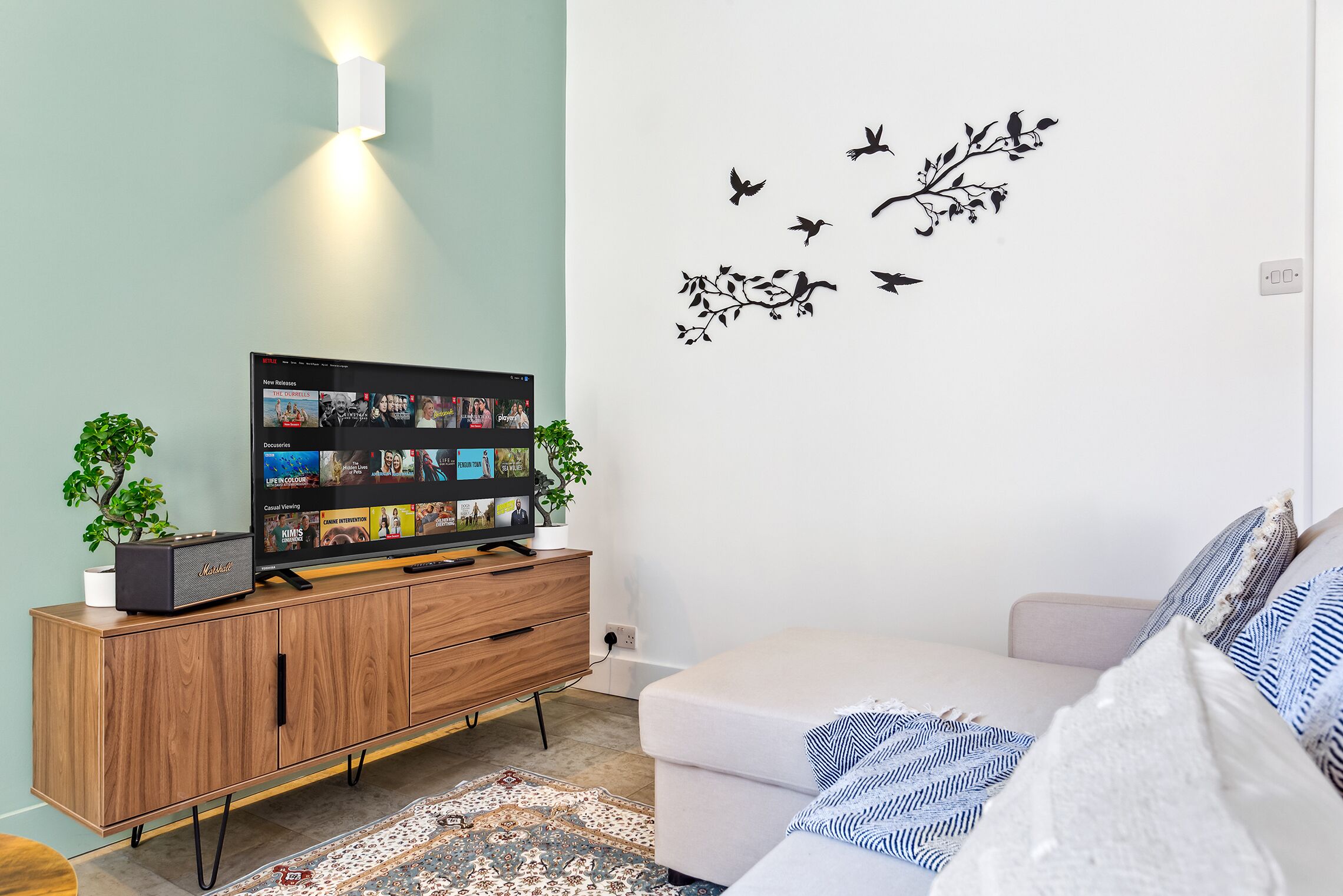 Ett vardagsrum med en platt-TV, ett träskåp, två krukväxter, en vit soffa och en väggdekor med fåglar.