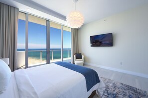 Ein Hotelzimmer mit einem großen Bett, einem Fernseher, einem Sessel und Blick auf den Ozean.
