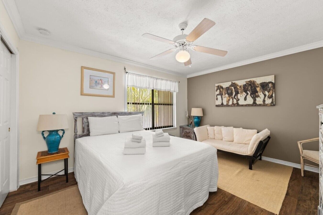 Quarto com cama, ventoinha no teto, janela com cortinas, sofá, poltrona e um quadro na parede.