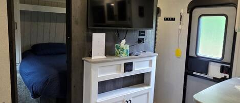 A compact RV interior with a bed, a white entertainment unit, and a small window.