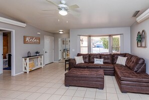 A spacious living room with a large sectional sofa, a ceiling fan, and a view through large windows. - Highlander Retreat - Pool, Pool Table, Comfort (Lake Havasu City)