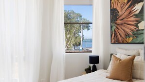 A bedroom with a bed, a nightstand, a lamp, and a view of trees and water through a window.
