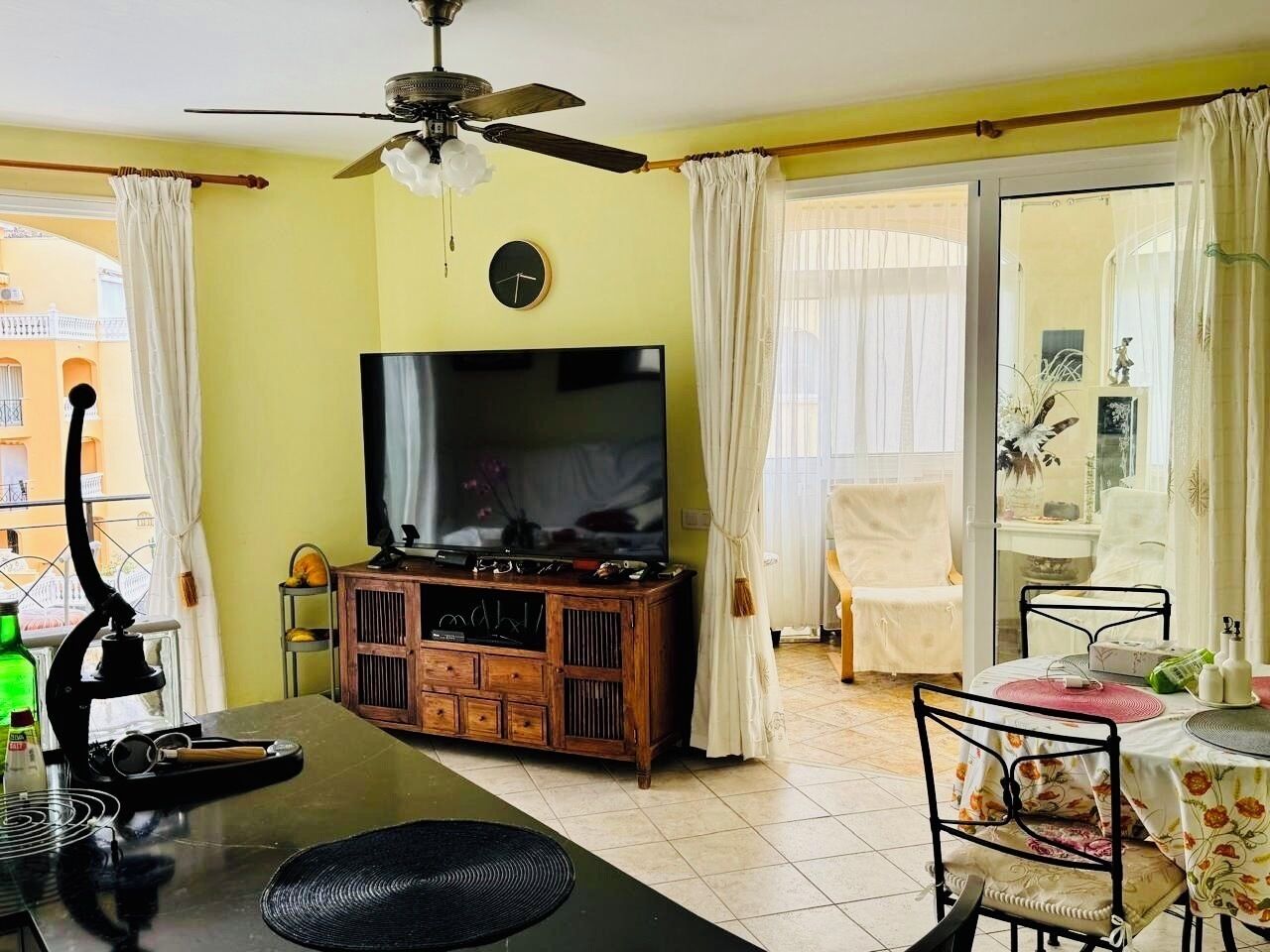 Ein Wohnzimmer mit Fernseher, Holzschrank, Esstisch, Stühlen und Deckenventilator.