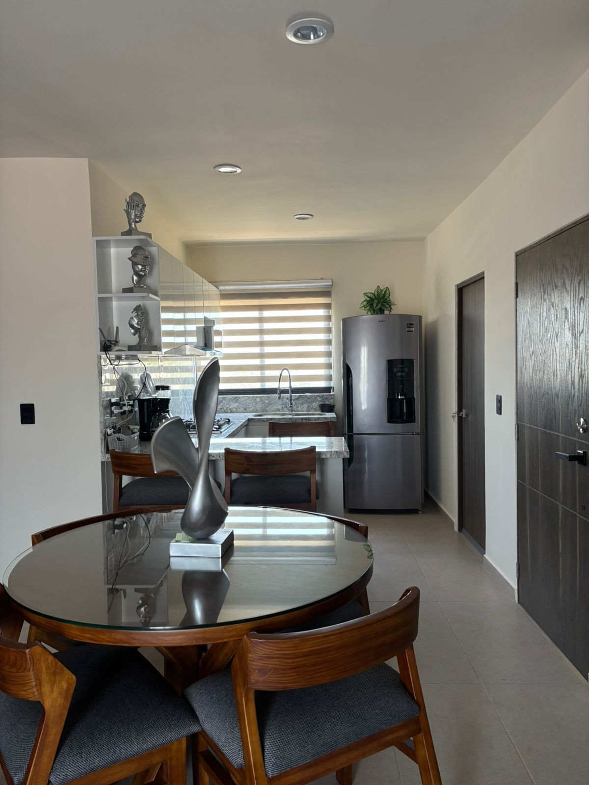 Una cocina moderna con comedor, que incluye una mesa redonda de cristal, sillas de madera y un refrigerador de acero inoxidable.