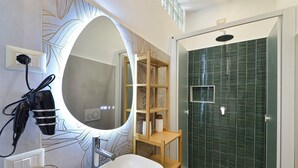 Un baño moderno con un espejo ovalado grande, un lavabo blanco, un estante de madera, una ducha con azulejos verdes y un perchero.
