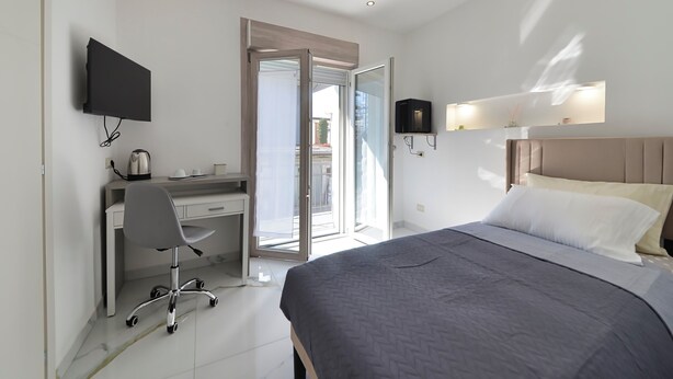 Habitación de hotel con cama, escritorio, silla, televisión y puerta corrediza al balcón.