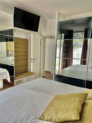 A hotel room with a bed, a seating area with a sofa and a chair, a television mounted on the wall, and a large mirror reflecting the room.
