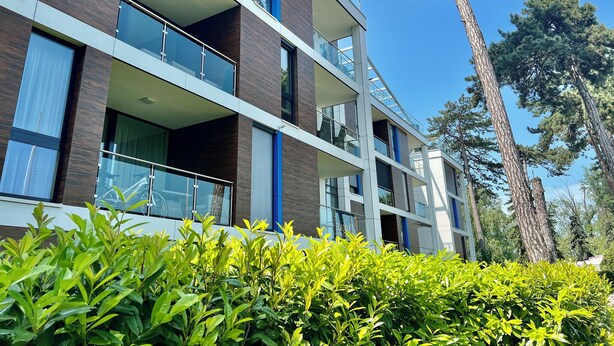 Un immeuble à plusieurs étages, doté de balcons, de grandes fenêtres et entouré d’un paysage verdoyant.