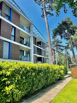 A modern multi-story building with balconies, a clear sky, and surrounding greenery.