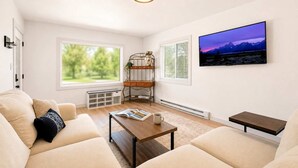 Un salón con sofá, mesa de centro y estantería. Hay dos ventanas, una con persianas y otra con vistas a zonas verdes. Un cuadro cuelga en la pared, y se ve una lámpara de techo.
