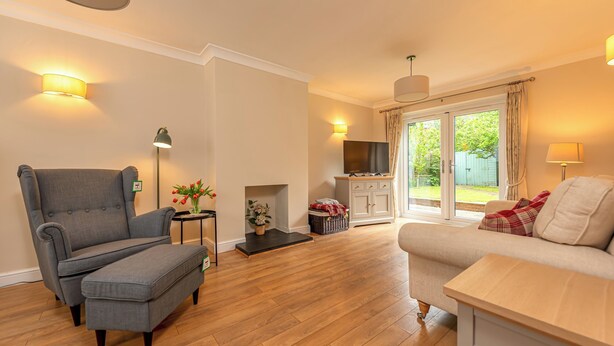 Een woonkamer met een grijze fauteuil, voetenbankje en een bank met een tafel. Er is een open haard, een televisie en uitzicht op de omgeving.