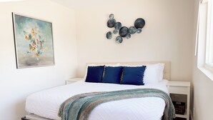 Una cama bien hecha con edredón blanco y manta azul, una obra de arte abstracto colgada en la pared y una mesita de noche con una cesta.