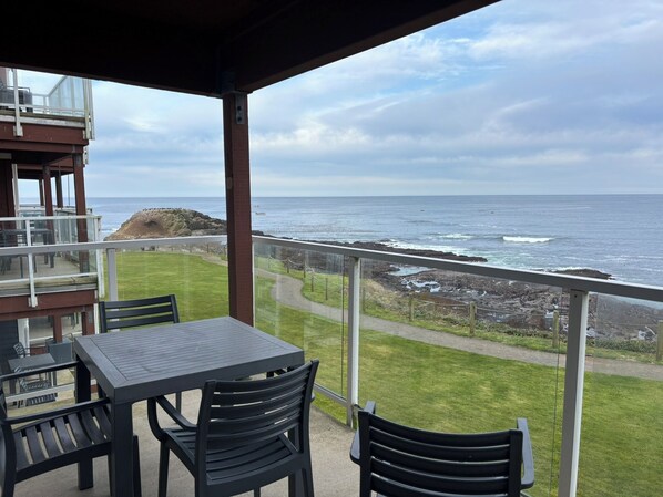 A balcony with a table and chairs overlooking a rocky coastline and the sea. - Charming 2-bedroom condo in Depoe Bay with ocean views (Depoe Bay)
