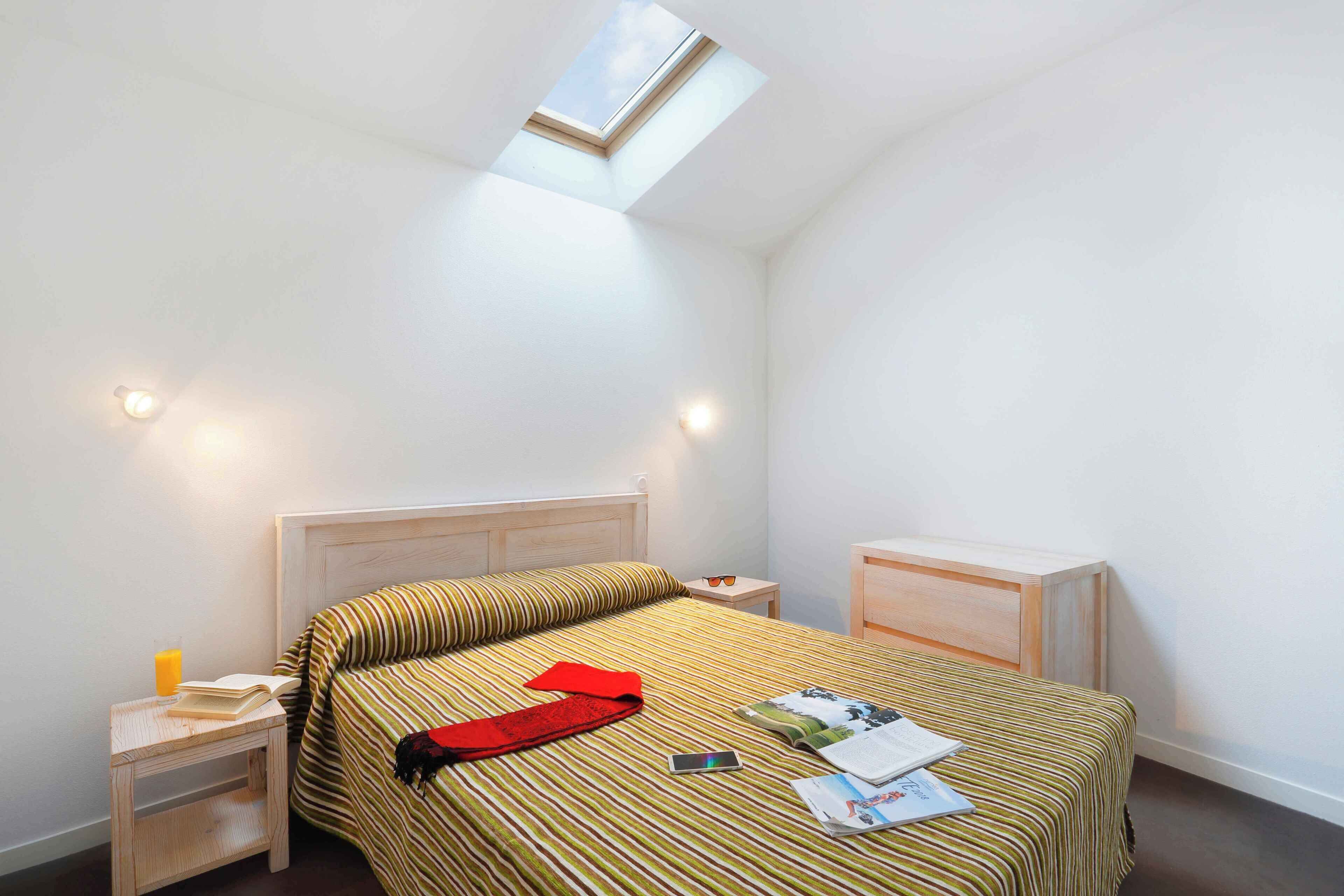 Un dormitorio con tragaluces, una cama con ropa de cama a rayas, una mesita de noche con un libro y gafas, y una mesita de noche con un vaso de jugo.