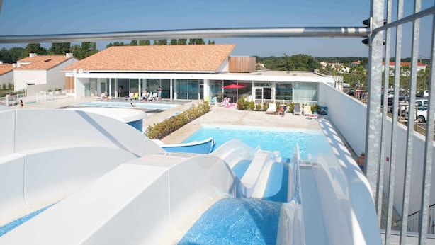 Parque aquático com várias piscinas e toboáguas, área coberta com assentos e um edifício com grandes janelas.