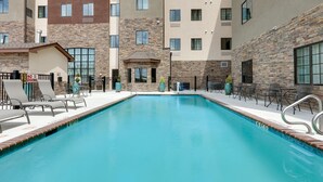 Une piscine entourée de chaises longues et agrémentée d’un panneau indiquant qu’il est interdit de plonger. Des immeubles à plusieurs étages aux façades en pierre et en brique l’entourent.