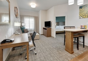 A hotel room with a desk, chair, bed, TV, and a painting on the wall.