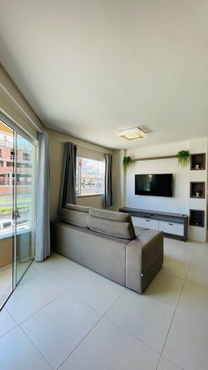 Ein modernes Wohnzimmer mit einer großen Couch, einem flachbildfernseher an der Wand und einer Glastür zum Außenbalkon.