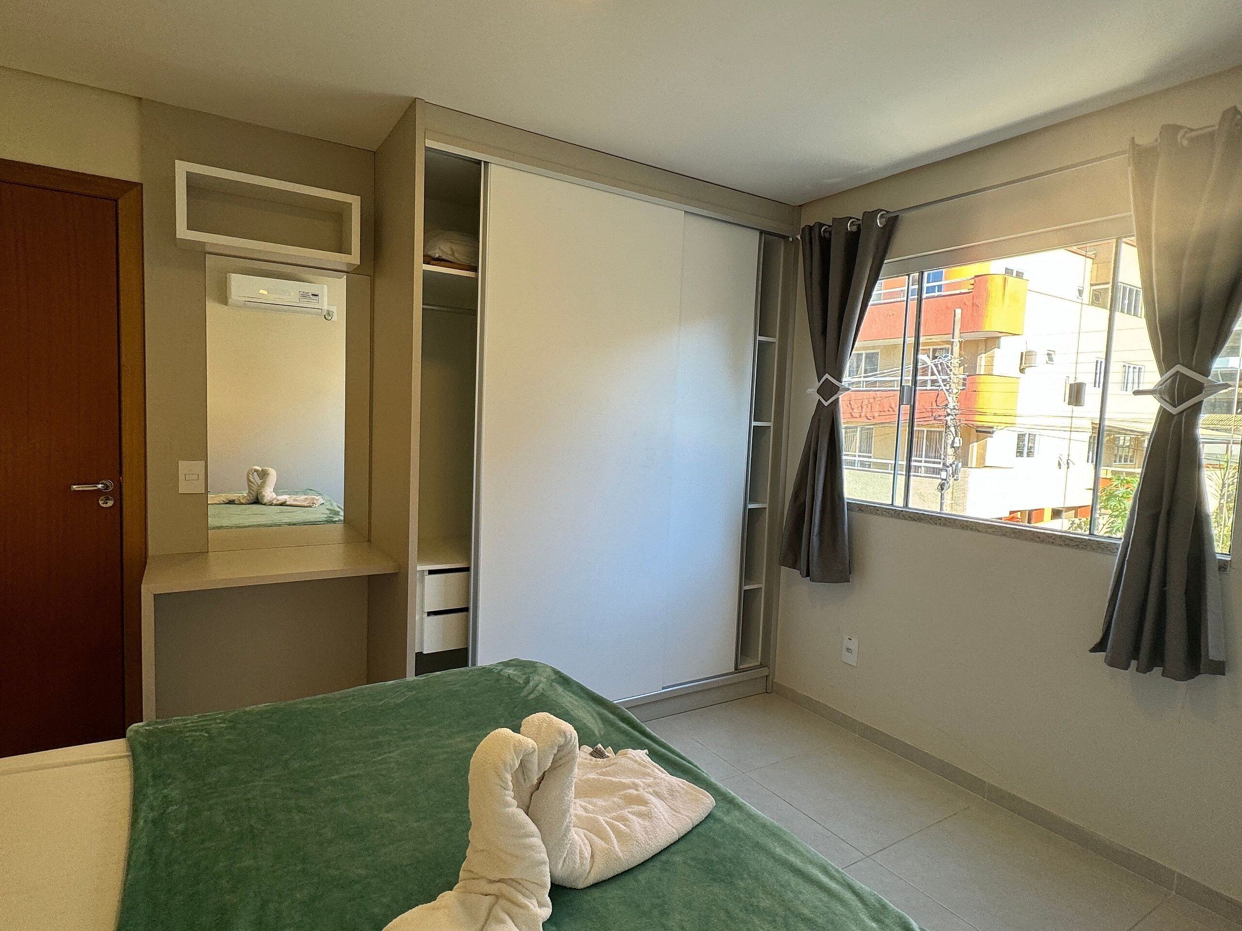 A hotel room with a bed, a green bedspread, a white towel, a wardrobe, and a window with curtains.