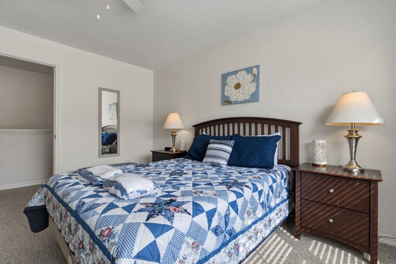 Un dormitorio con una cama, dos mesitas de noche, un tocador, un espejo, un adorno floral en la pared y una puerta.