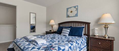 Un dormitorio con una cama, dos mesitas de noche, un tocador, un espejo, un adorno floral en la pared y una puerta.