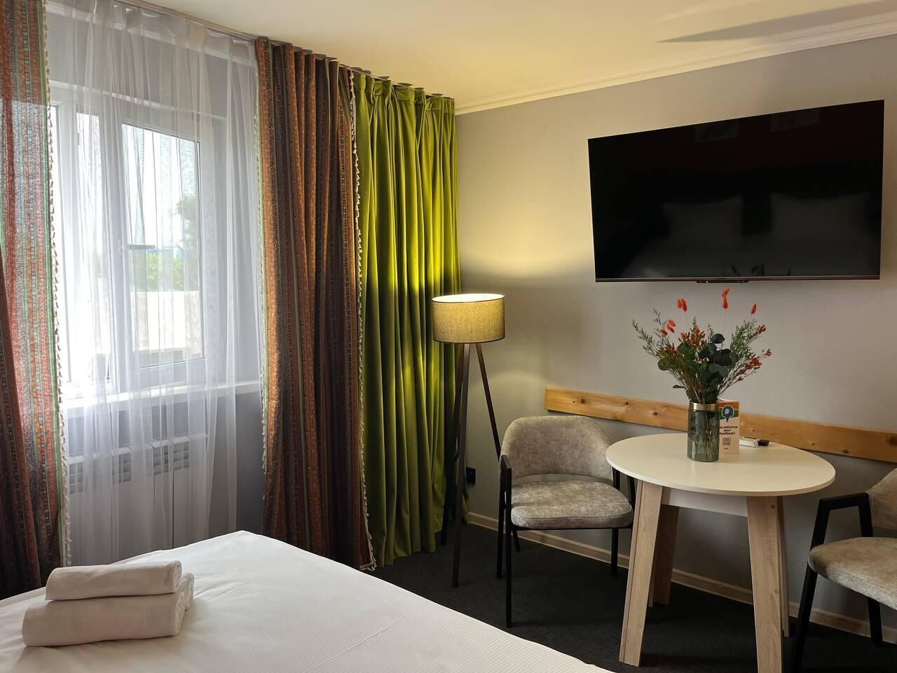 Une chambre d’hôtel avec un lit, un bureau, deux chaises, une télévision et une fenêtre avec des rideaux.