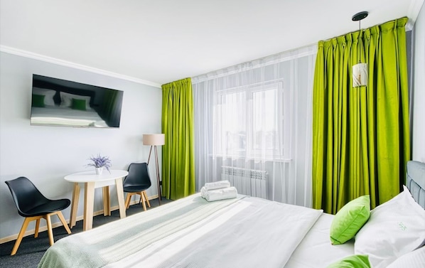 A hotel room with a bed, a desk with chairs, a small table with a plant, and a window with sheer curtains. - The Apartamens 95 (Almaty)
