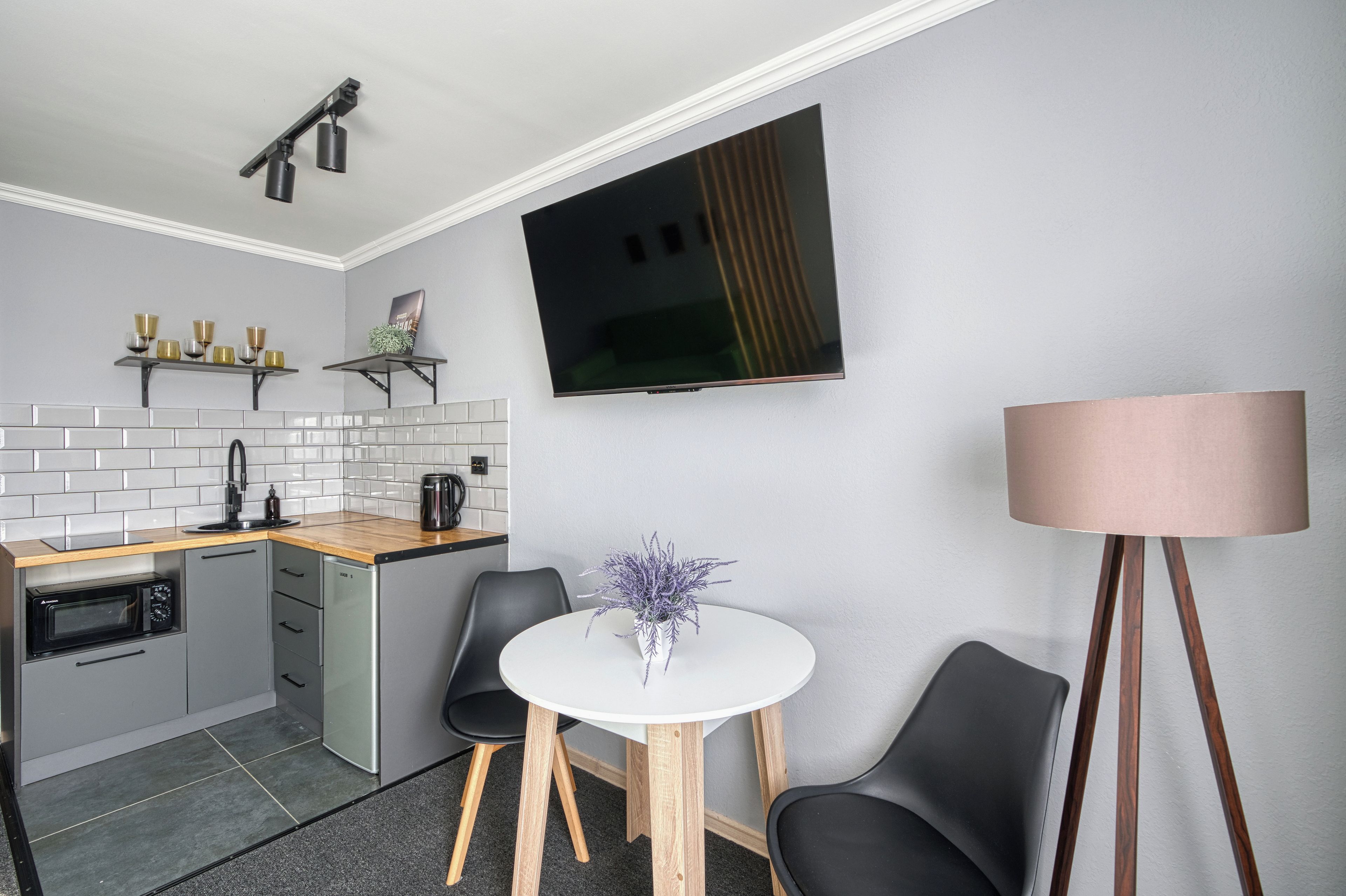 Een praktische keuken met een magnetron, waterkoker en een klein spoelbakje. Er staat een witte tafel met stoelen bij, en een potplant. Een flatscreen televisie is aan de muur bevestigd.