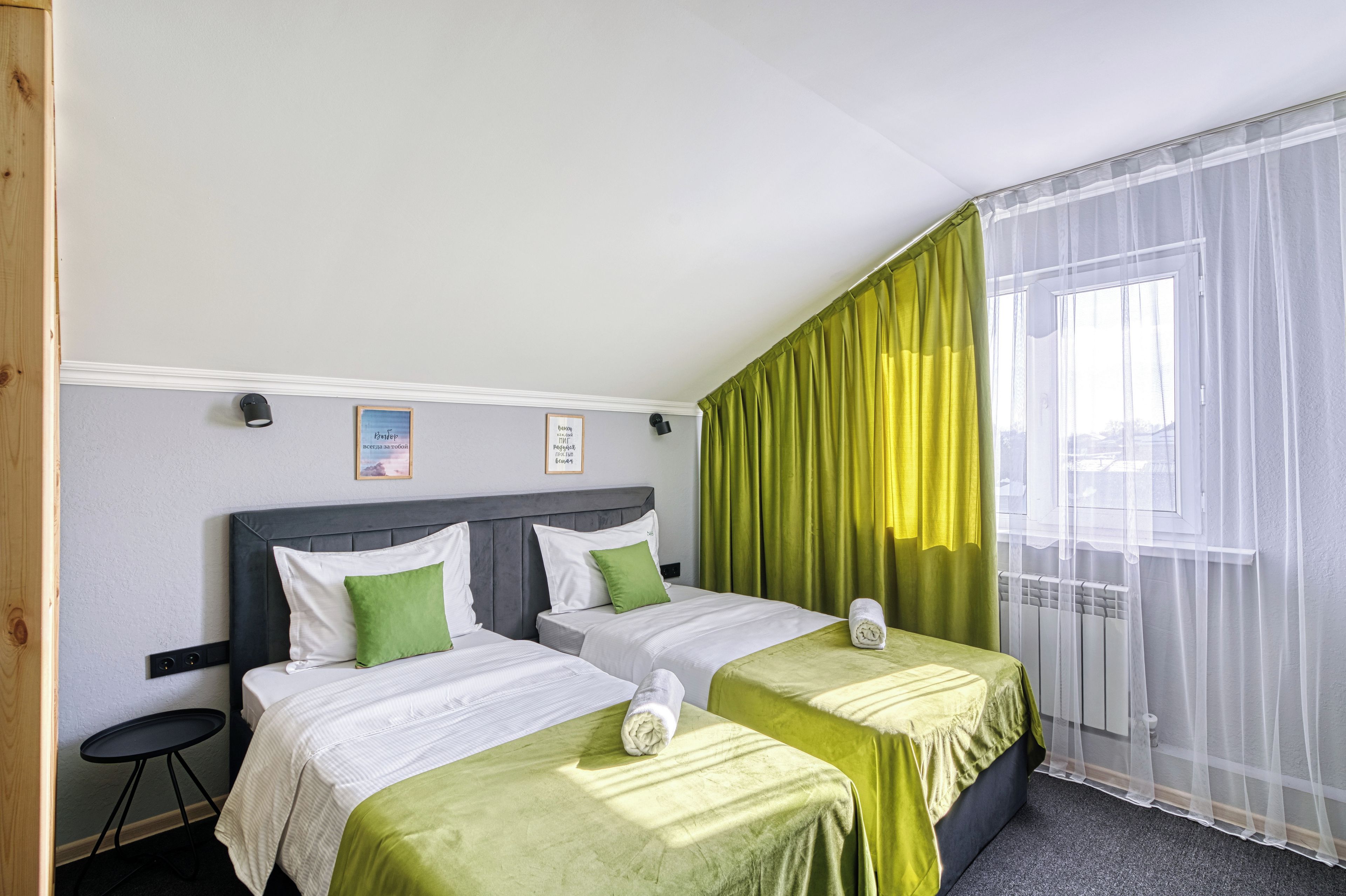 A hotel room with two beds, a small table, a window with sheer curtains, and a radiator.