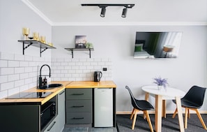 Cocina compacta con lavamanos, estufa y un refrigerador pequeño. Hay un comedor con mesa y sillas, y un televisor de pantalla plana montado en la pared.
