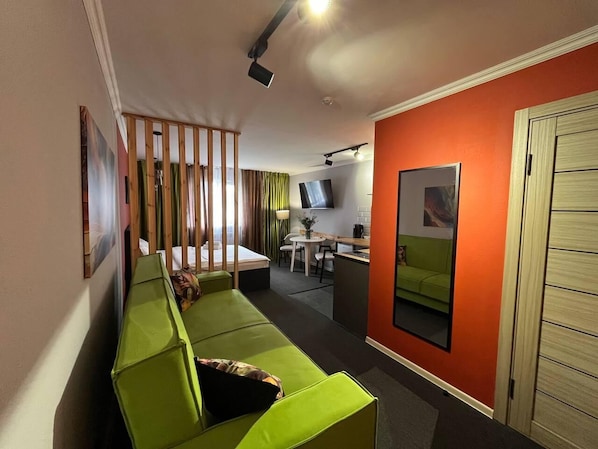 Una habitación compacta con sofá verde, una pequeña mesa de comedor, televisor y kitchenette.