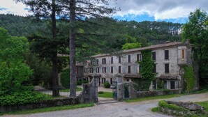 Un edificio rústico de piedra con varias ventanas y una entrada porticada, rodeado de exuberante vegetación y árboles.