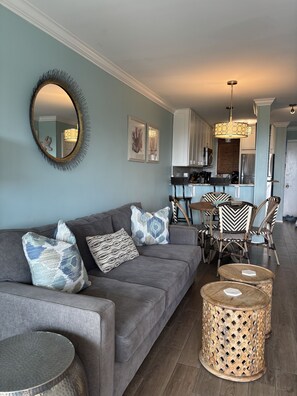 Ein Wohnzimmer mit grauem Sofa, gemusterten Kissen, einem runden Spiegel und einem Essbereich mit Korbstühlen und einem Holztisch.