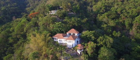 Una casa grande rodeada de bosque denso, con varias edificaciones y un sendero visible.